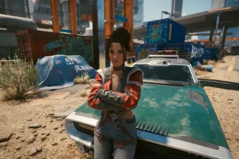 Panam z Cyberpunk 2077 stoi obok swojego zielonego pojazdu, z rękami skrzyżowanymi na piersi, w postapokaliptycznym otoczeniu.