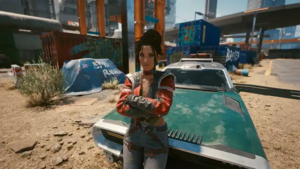 Panam z Cyberpunk 2077 stoi obok swojego zielonego pojazdu, z rękami skrzyżowanymi na piersi, w postapokaliptycznym otoczeniu.